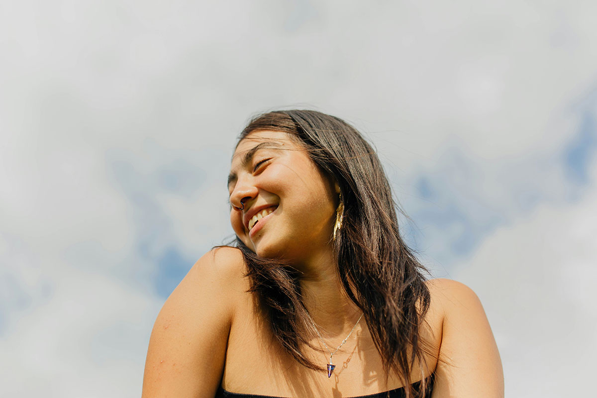 Young woman smiling in front of a blue sky filled with puffy white clouds.